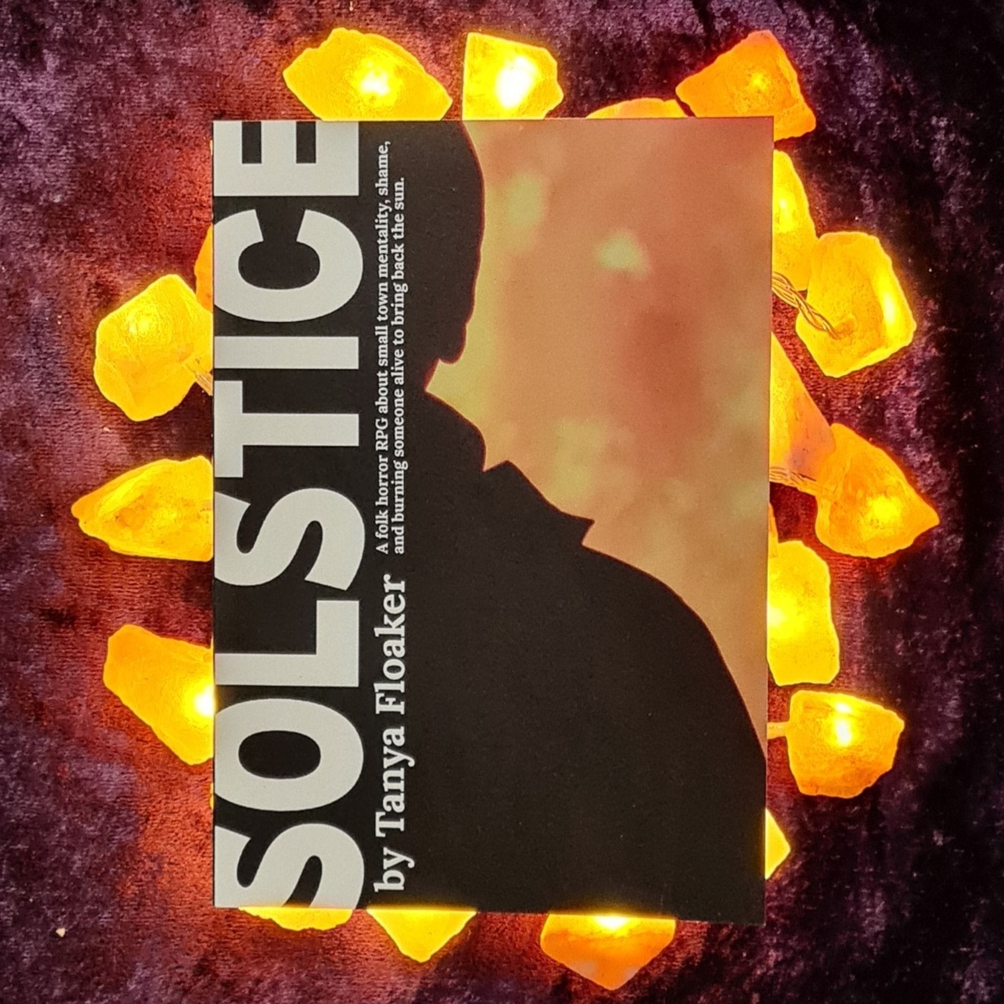 A copy of the Solstice RPG zine sits on top of some amber-glowing rock crystal fairy lights, all sat on a purple velvet cloth. The zine has the half sillouette of a person on cut down the middle on the left-and spine. To their right rages fire. In their sillouette is a banner reading "SOLSTICE / by Tanya Floaker / A folk horror RPG about small town mentality, shame, and burning someone alive to bring back the sun."