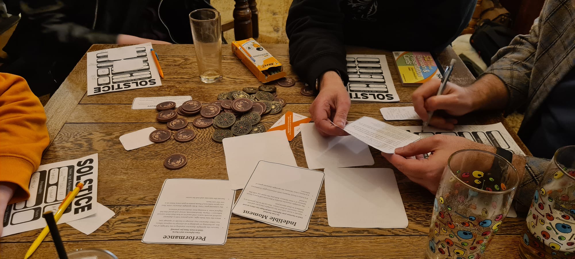 People sat at a pub table playing Solstice. The table is strewn with tokens, character sheets, game components cut from the book, and glasses. 