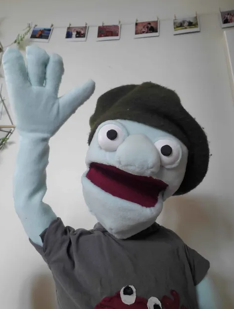 A puppet with blue felt skin and a beret smiles at the camera, raising one arm.