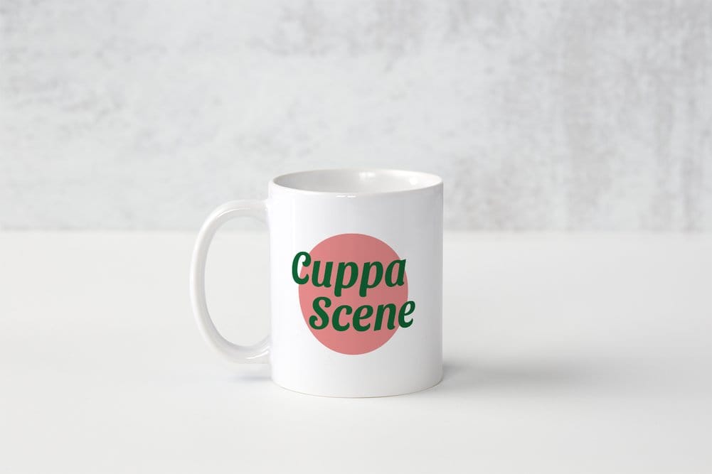 A Mum Chums white mug sat on a grey background. It has a salmon pink circle with the words "Cuppa Scene" over the top in green. 