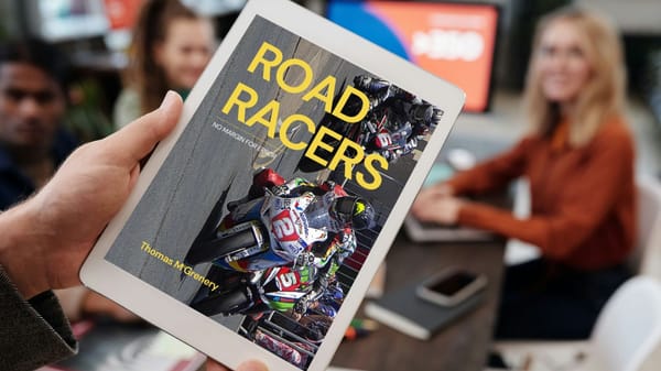 Mockup of Road Racers cover on a tablet being held by someone addressing a group of people seated at a table. 