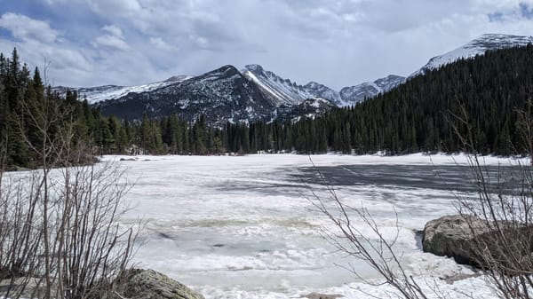 A photo of Bear Lake in Rocky Mountain National Park. a mostly frozen lake foregrounds green pines and distant, snowy peaks.