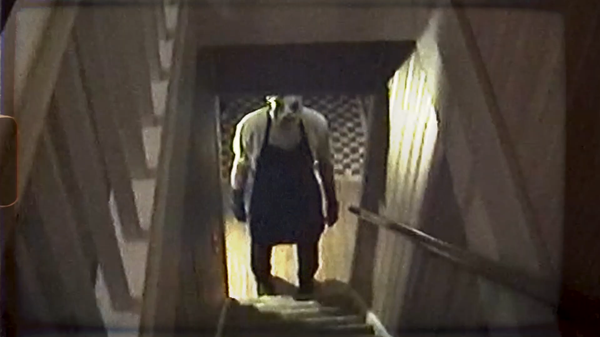 A person in a blck apron and deformed mask stands ominously at the bottom of a staircase.