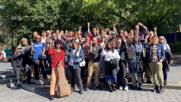 Dozens of workers stand in a park, fists raised in solidarity. Bright afternoon light illuminates green trees.