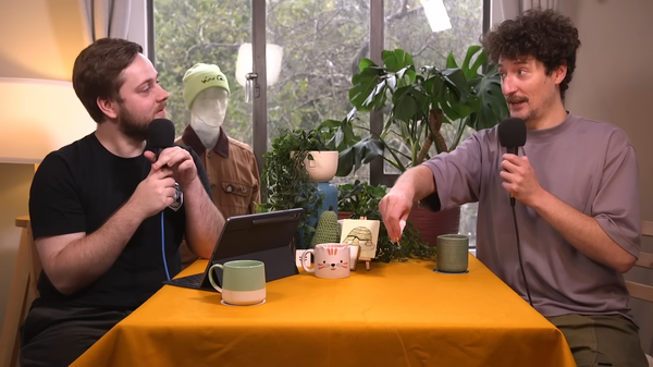 Tom Brewster and Matt Lees sit at a yellow table holding microphones. Mugs, plants and a window fill the small room.