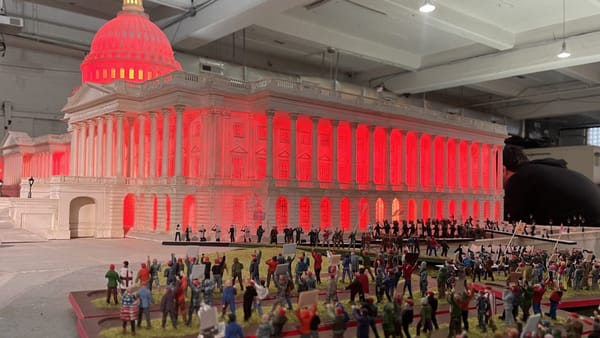 A flood of miniature MAGA figurines surrounding a model of the US Capitol Building lit in red