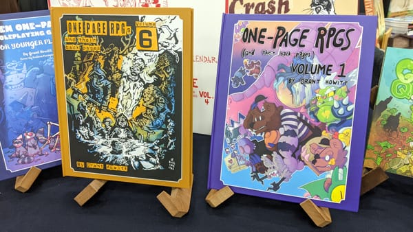 Volumes 1 and 6 of One-Page RPGs on wooden book plinths at GAMA 2025. Other books join it on a black display table.