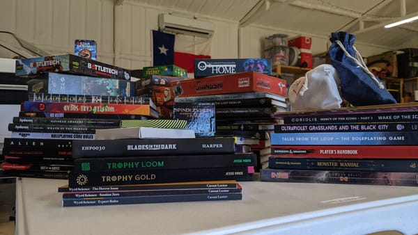 Several stacks of tabletop RPG books and board games on a white table. A boat and Texas flag seen in the background.